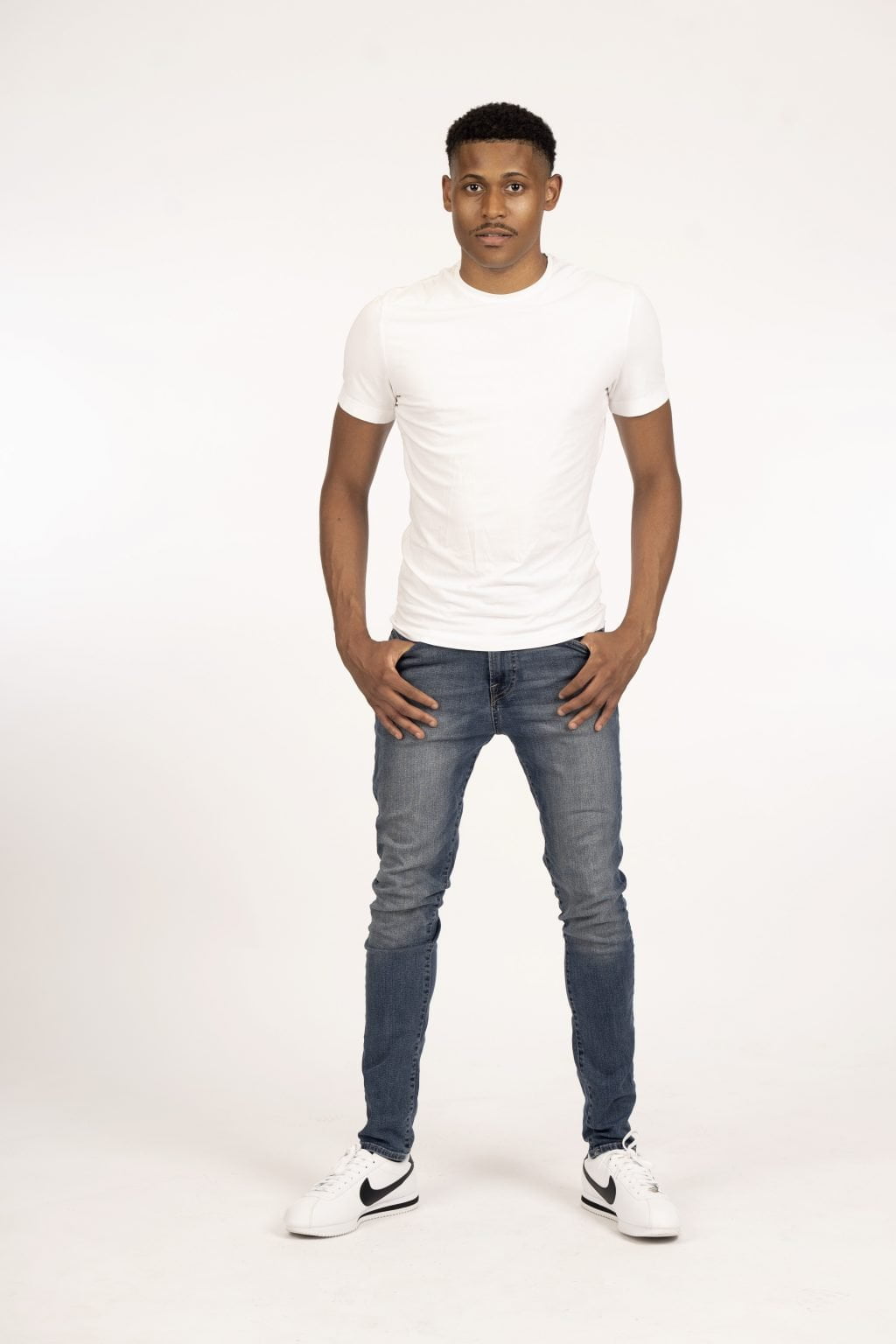 Full body shot of Tyler Jurelle in a white shirt, blue jeans, and white Nike Cortez with a black swoosh logo, standing against a white backdrop.