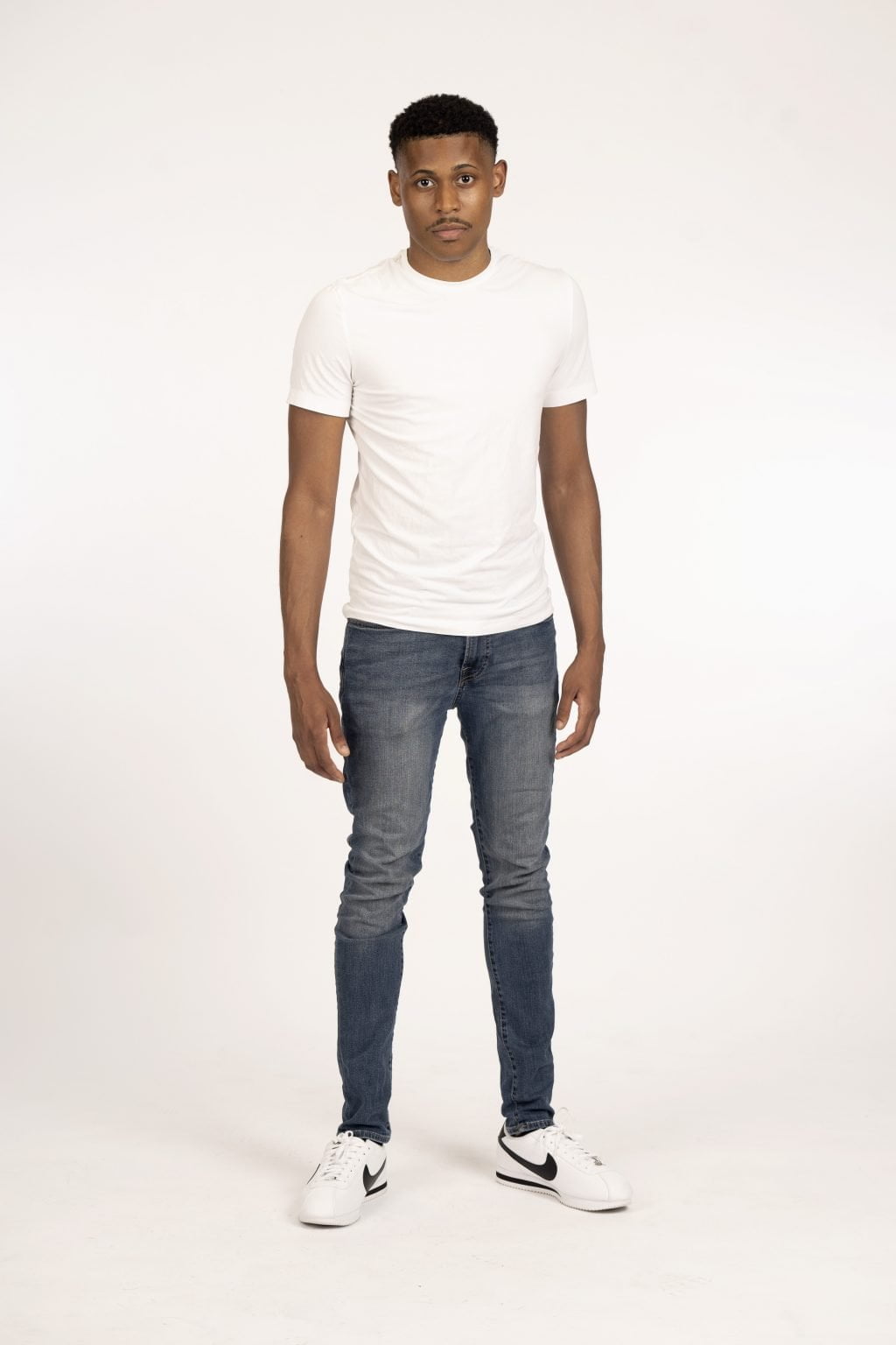 Tyler Jurelle showcasing timeless style in a full body shot, wearing a white shirt, blue jeans, and Nike Cortez shoes, standing in front of a white backdrop.