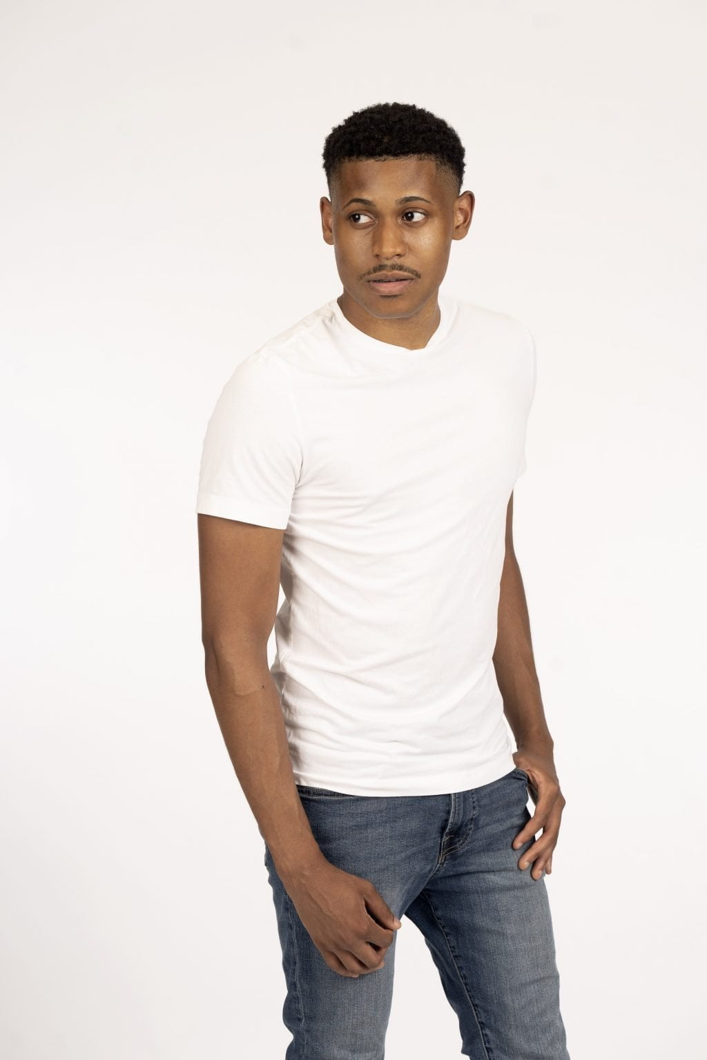Tyler Jurelle in a 3/4 body side profile, wearing blue jeans and a white shirt, looking away from the camera against a white backdrop.