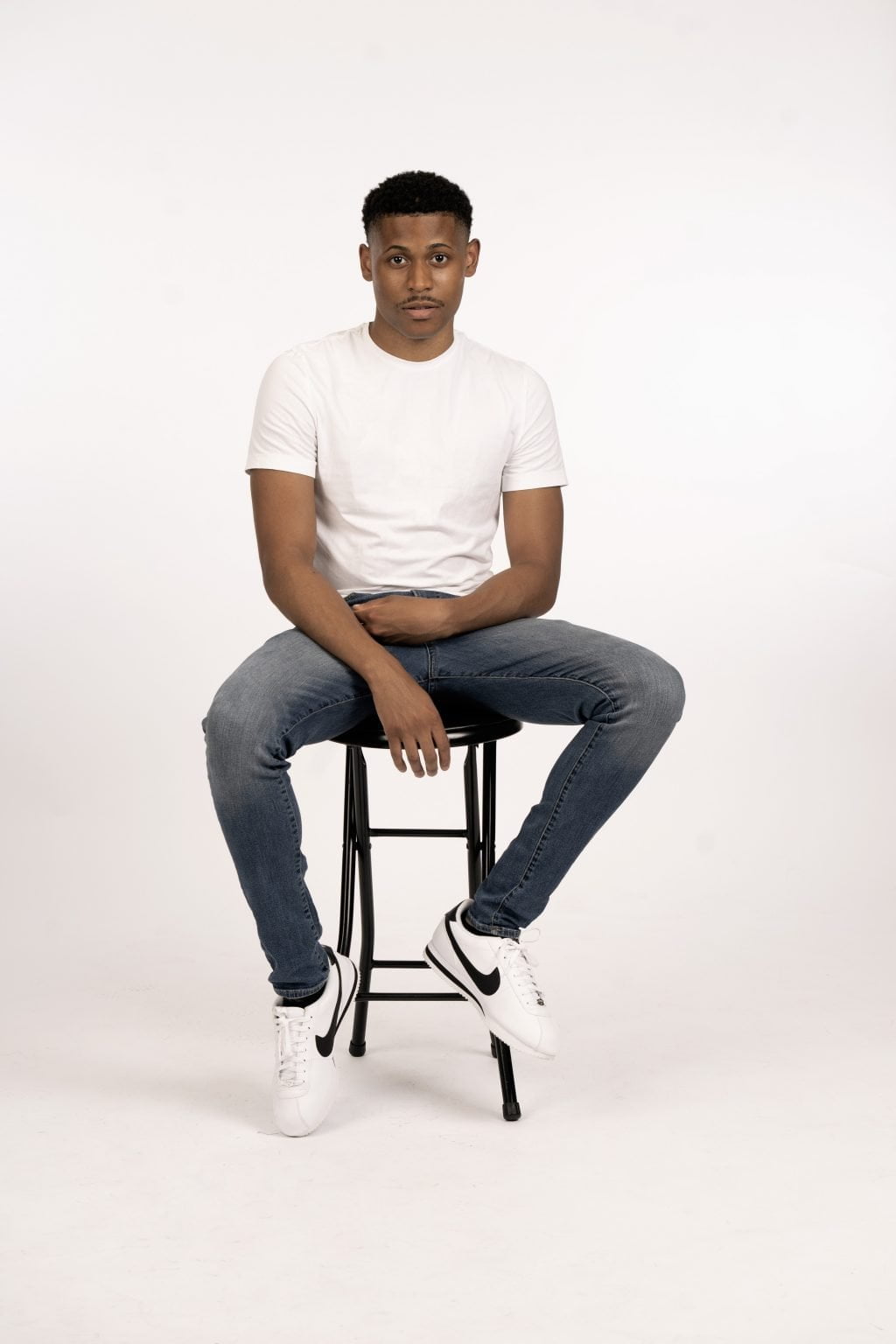 Sitting down on a stool, front-facing full body shot, dressed in a white shirt, blue jeans, and Nike Cortez shoes, set against a white backdrop.