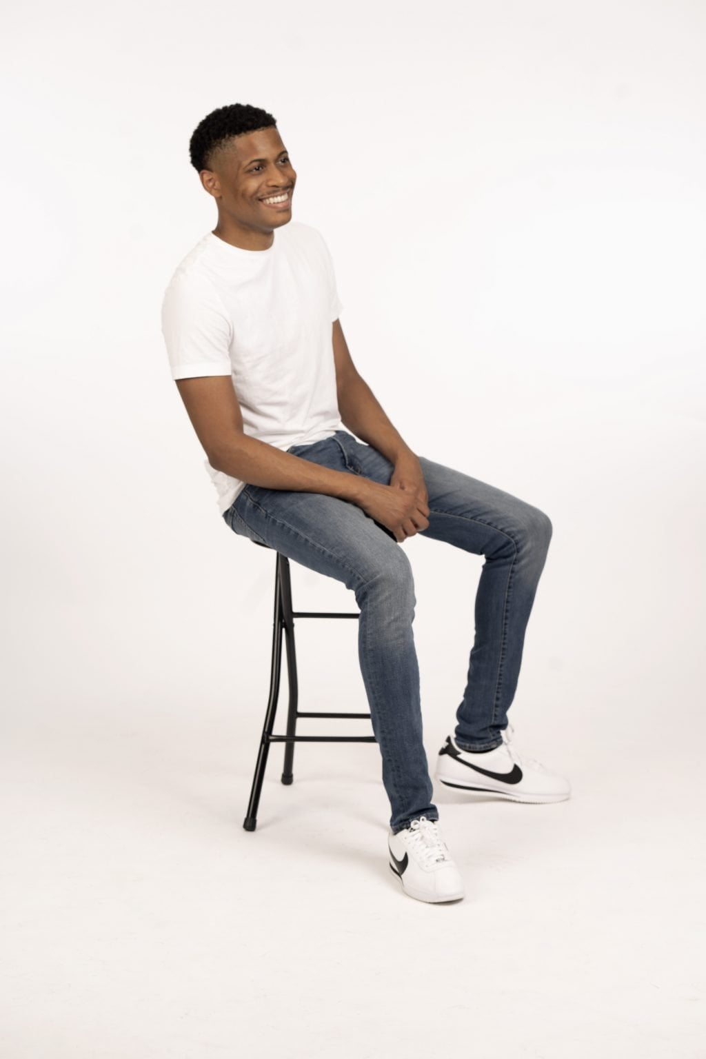Smiling and looking away from the camera at a 45-degree angle, Tyler Jurelle sits on a stool in a white shirt and blue jeans, wearing Nike Cortez shoes.