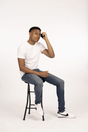 Tyler Jurelle is holding a brush, brushing his hair, sitting on a stool, dressed in a white shirt, blue jeans, and Nike Cortez shoes on a white backdrop.