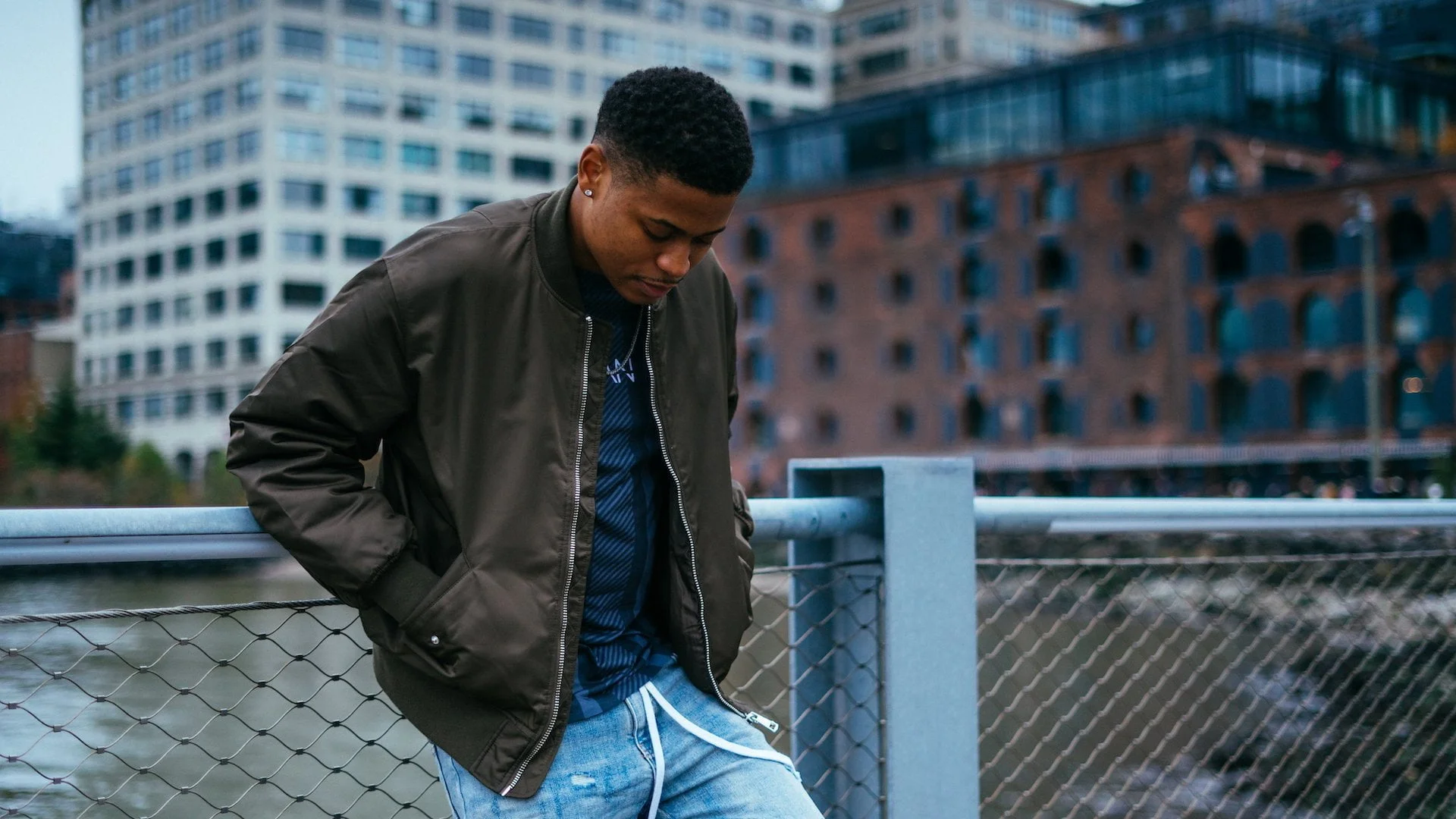 At Brooklyn Bridge Park, Tyler Jurelle strikes a pose in an olive bomber jacket and jeans. Book him for his modeling services.