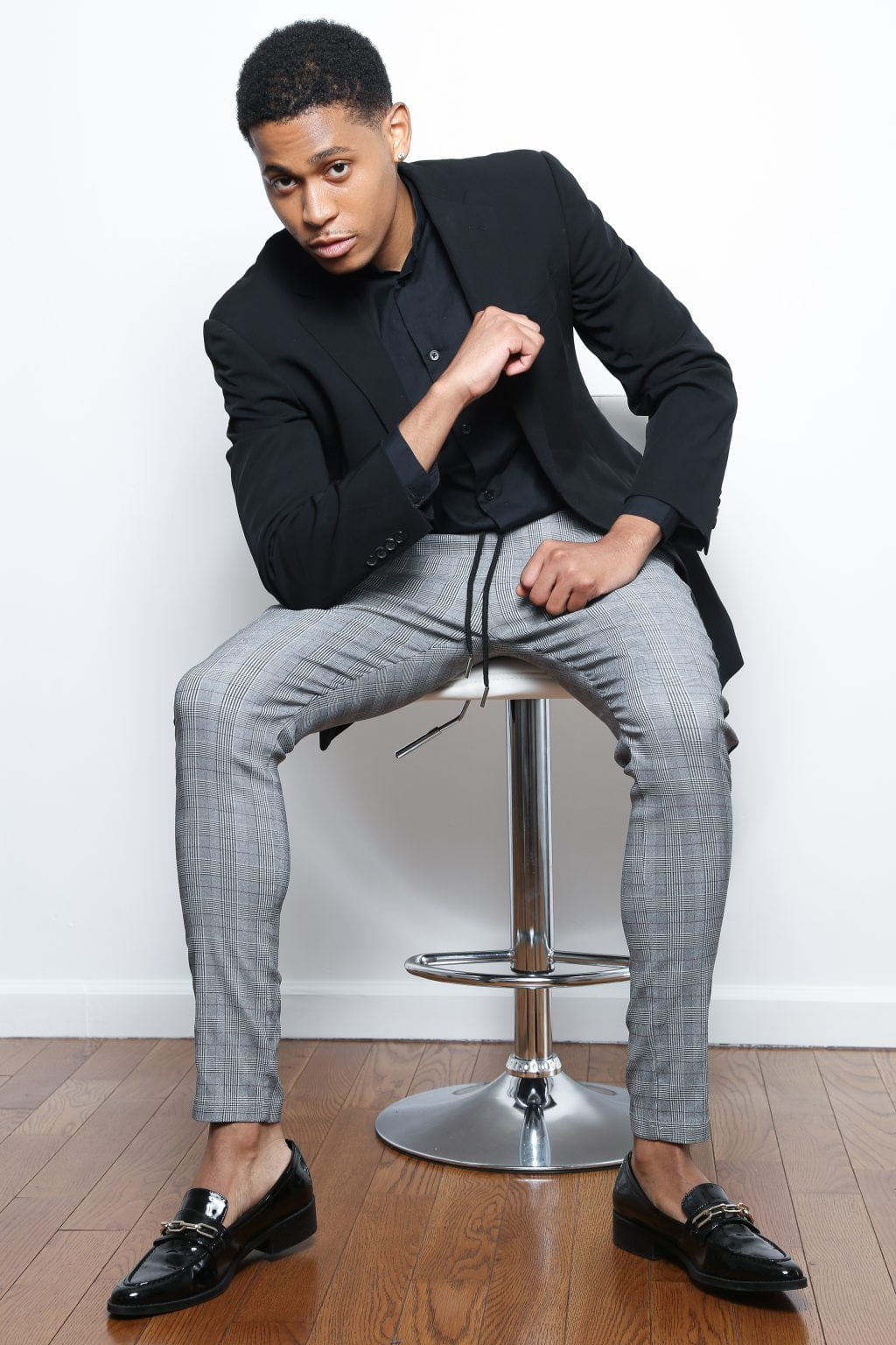 Tyler Jurelle sits poised in Deception Toronto’s Gray Miami Plaid Trousers and a black blazer, blending sophistication with modern fashion.