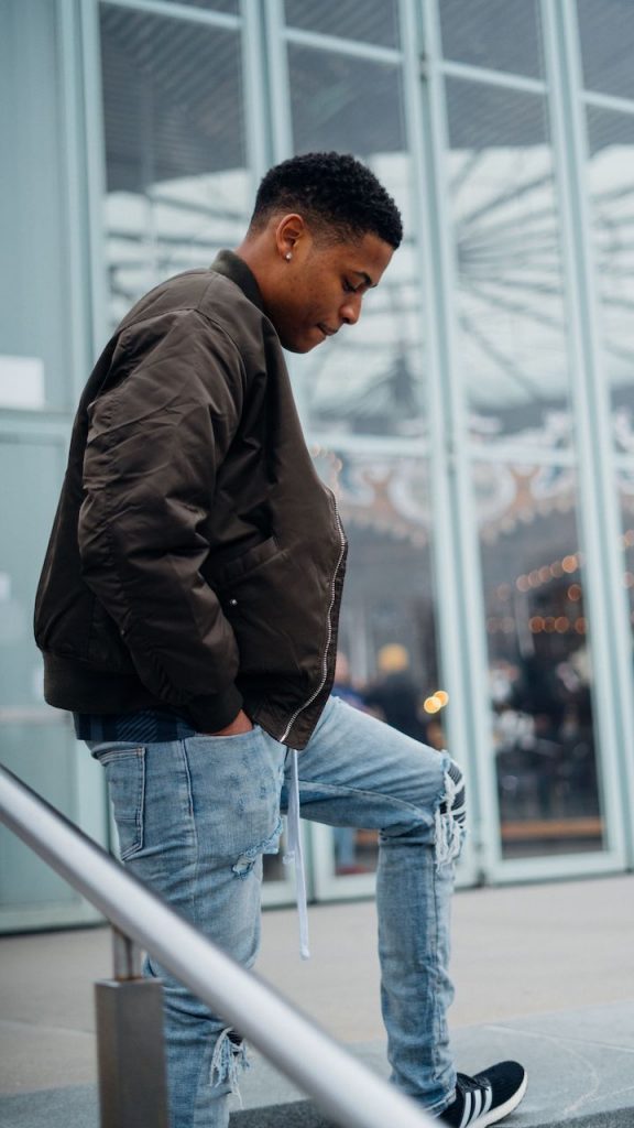 Displaying Biker Patch Denim and an olive green bomber jacket at Brooklyn Bridge Park, Tyler Jurelle stands with a relaxed posture.