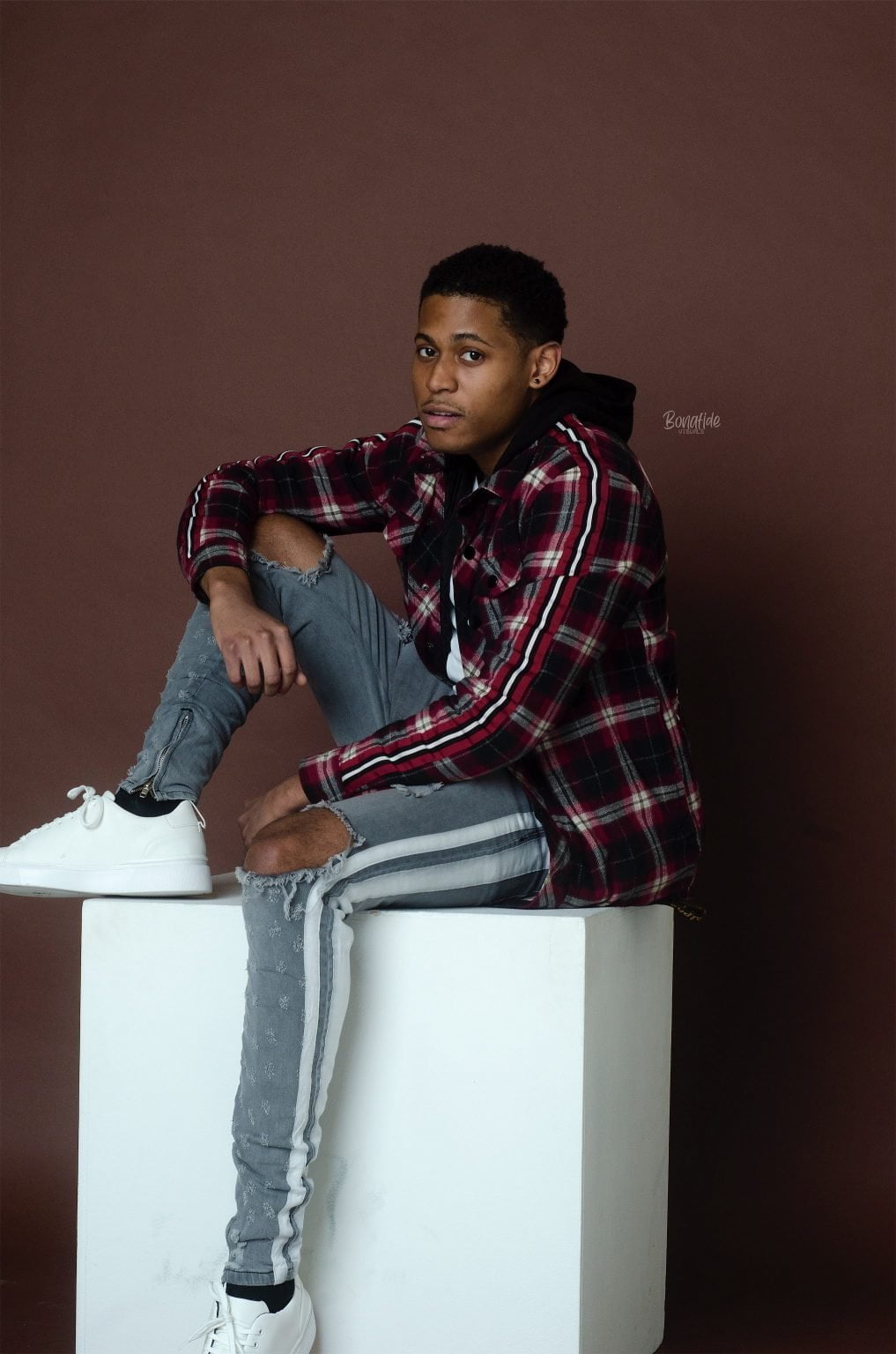 Tyler Jurelle is sitting on a cube platform in a full-body studio portrait, showcasing a relaxed yet poised look against a neutral backdrop.