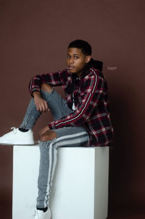 Tyler Jurelle is sitting on a cube platform in a full-body studio portrait, showcasing a relaxed yet poised look against a neutral backdrop.