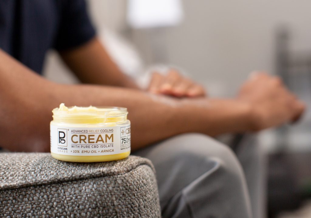 With an open jar of CBD cream in the foreground, Tyler Jurelle is applying Physicians Grade CBD cream with precision, showcasing its smooth texture.