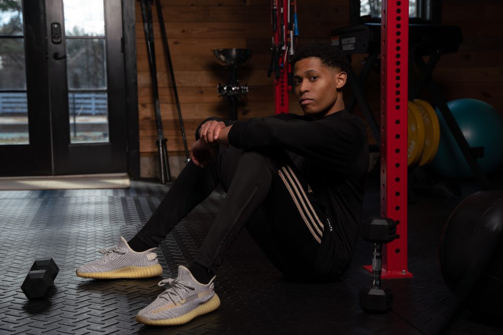 In a reflective gym moment, male model Tyler Jurelle is seated with dumbbells by the side, capturing a pause in training.
