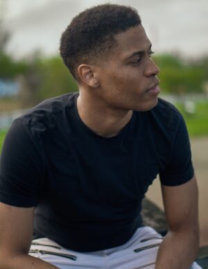 A close-up of Tyler Jurelle sitting on a wooden post, dressed in a black t-shirt.