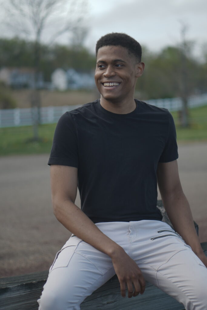 Tyler Jurelle smiles while sitting on a wooden post, wearing a black t-shirt and khaki cargo jeans.