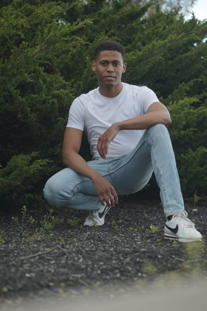 Posing in a deep squat, Tyler Jurelle showcases a casual outdoor look with a white t-shirt and jeans.