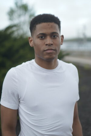 A clear headshot of Tyler Jurelle wearing a plain white t-shirt against a natural outdoor backdrop.