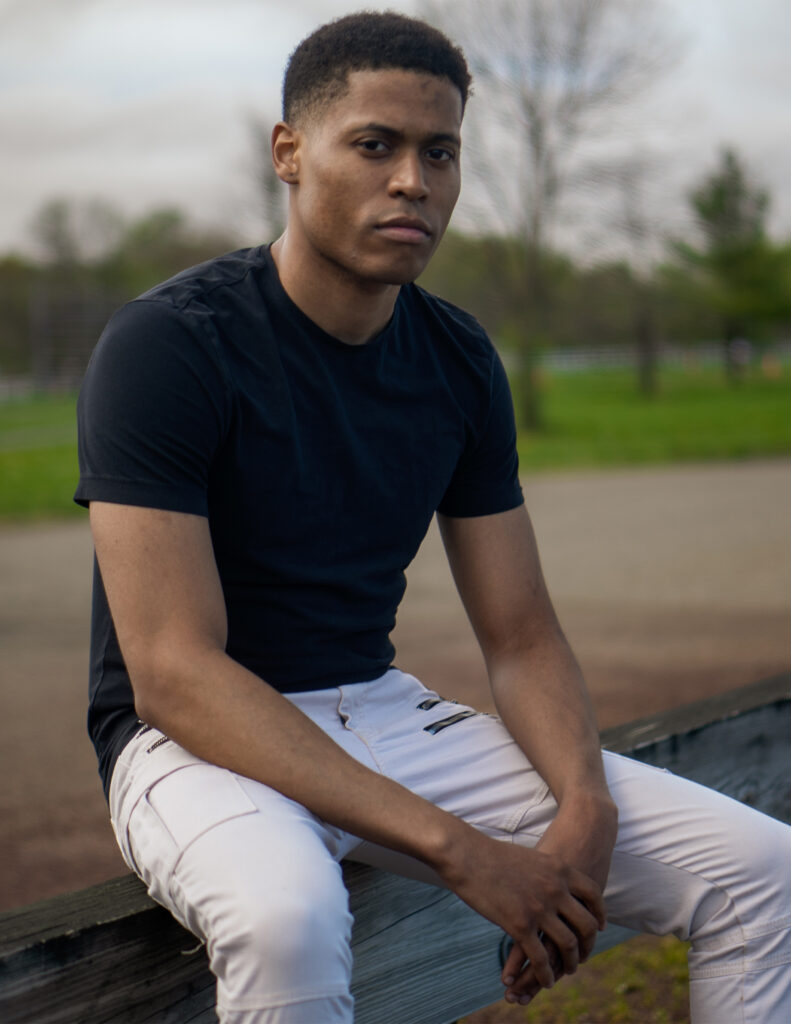 Tyler Jurelle sitting on a wooden post outdoors, in a black t-shirt and khaki cargo jeans.