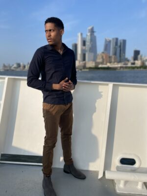Men's fashion creator Tyler Jurelle is standing on a boat with the NYC skyline in the background, dressed in quality after work attire.